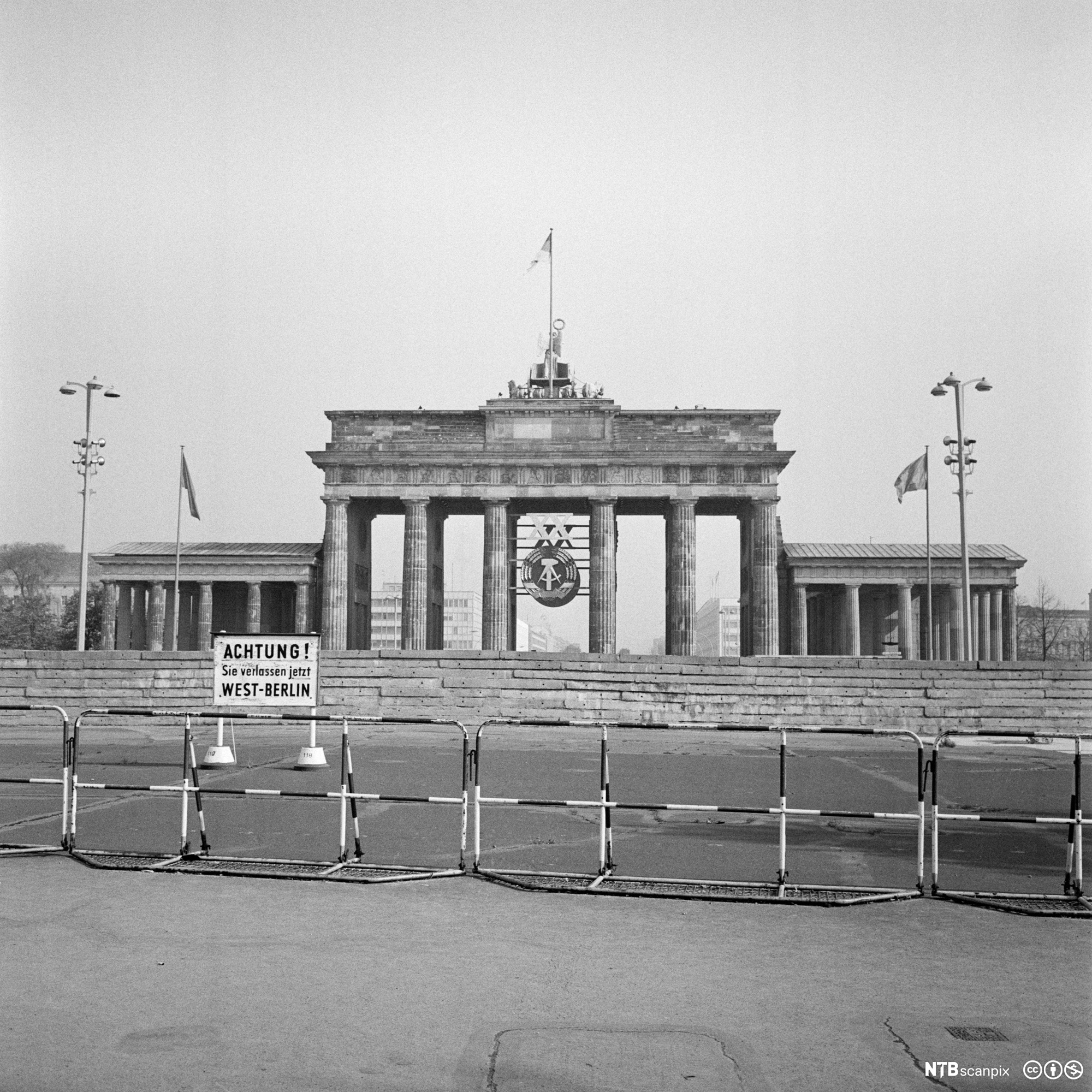 Brandenburger Tor fotografert fra Vest-Berlin i 1969. Fremst i bildet er det et enkelt gjerde, bak det et skilt der det på tysk står en advarsel om at man forlater Vest-Berlin. Deretter en høy mur foran selve Brandenburger Tor. Inni det historiske monumentet er det hengt opp et stort symbol for DDR-staten. Foto. 