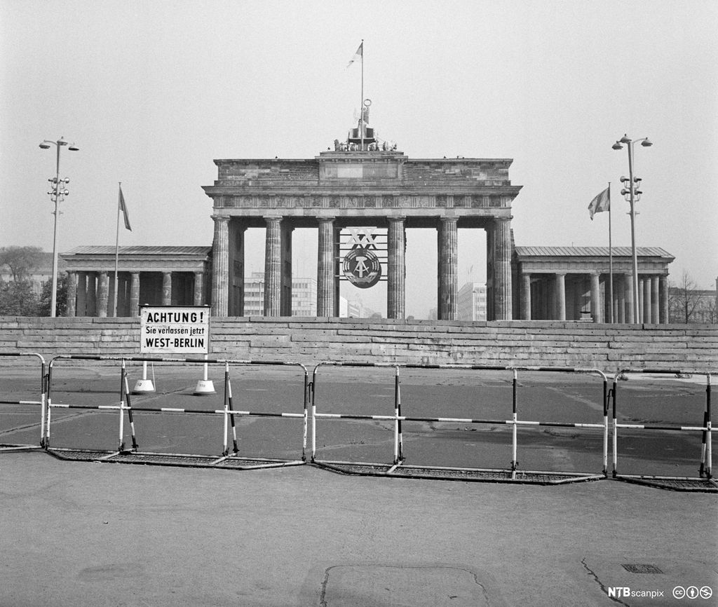 Brandenburger Tor fotografert fra Vest-Berlin i 1969. Fremst i bildet er det et enkelt gjerde, bak det et skilt der det på tysk står en advarsel om at man forlater Vest-Berlin. Deretter en høy mur foran selve Brandenburger Tor. Inni det historiske monumentet er det hengt opp et stort symbol for DDR-staten. Foto.