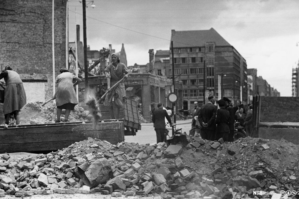 Nokre kvinner lempar grus og stein frå ei trekasse ned på gata. Nokre menneske står i ei gruppe bak ein stor haug  grus og stein. Foto. 