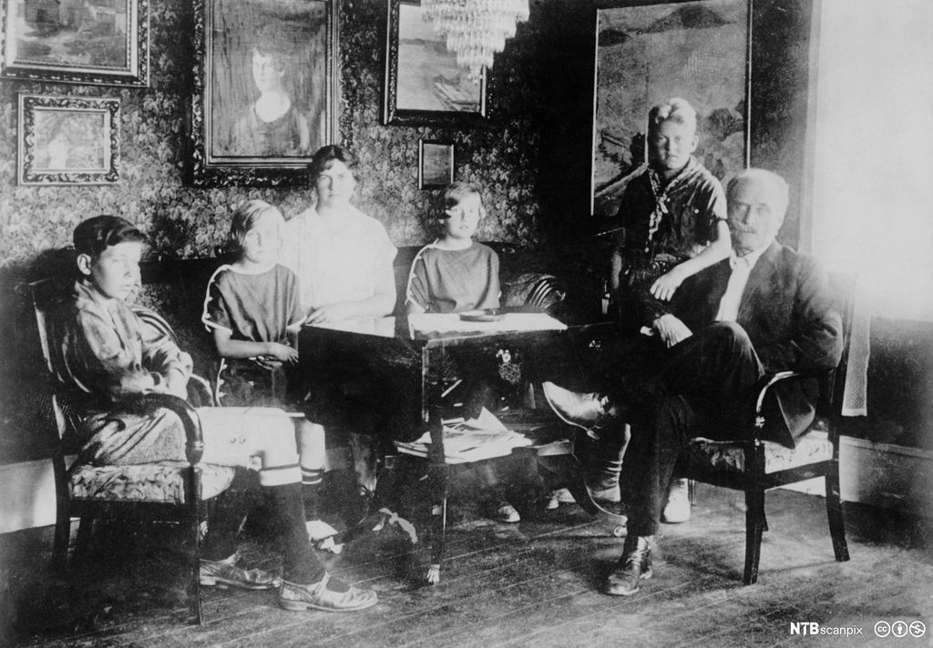 Knut Hamsun med kone og barn sitter rundt et bord i stua på Nørholmen. Fotografi.