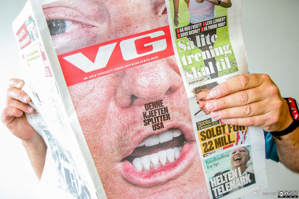 En person holder opp papirutgaven av VG fra 17. august 2017. Forsiden har et stort nærbilde av ansiktet til Donald Trump. Tittelen "Denne kjeften splitter USA" er plassert som en firkant mellom nesa og munnen. Foto.