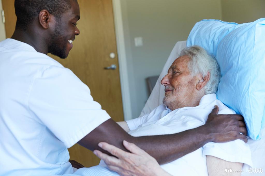 Mannlig helsefagarbeider holder armen på skulderen til eldre mannlig pasient som ligger i senga. Foto. 