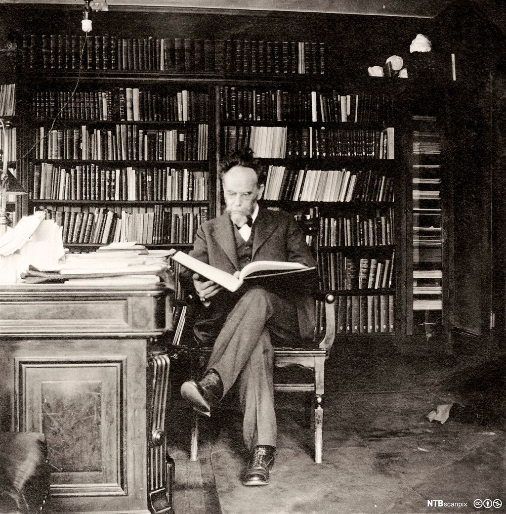 Portrett av den tysk-amerikanske antropologen Franz Boas i sitt arbeidsrom. Han sitter med en stor bok oppslått i fanget. Foto. 