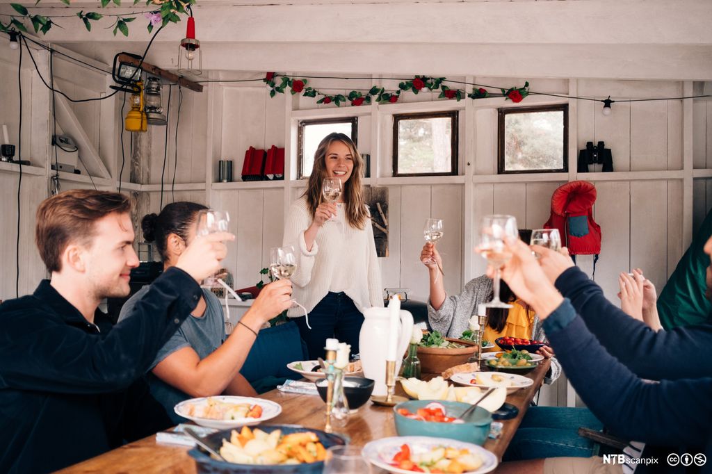 Flere personer rundt et bord dekket med mat, hever glassene og smiler. Foto.