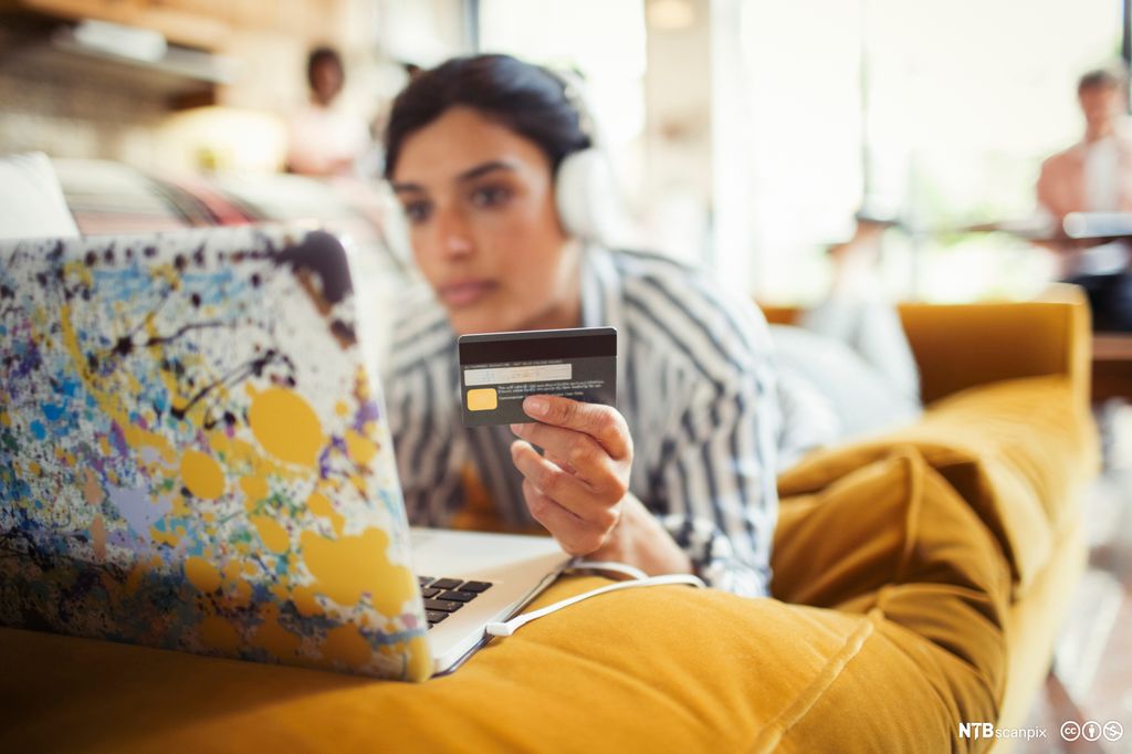 Ung kvinne med hovudtelefonar og kredittkort utfører netthandel på ein laptop. Foto.