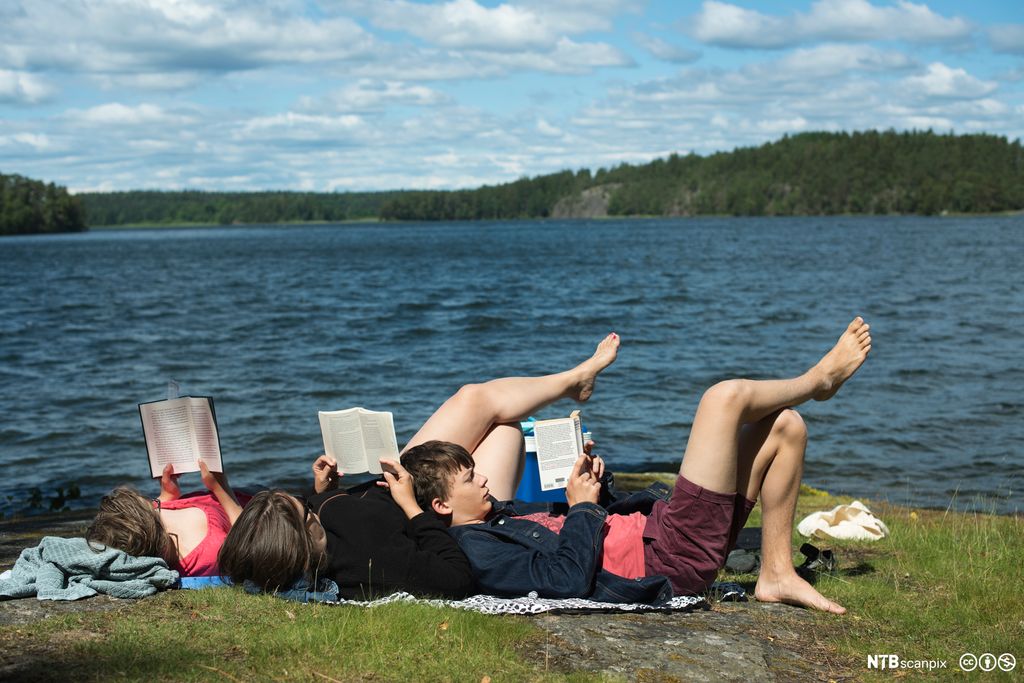 Tre personar ligg i graset ved ein innsjø og les. Det er sommar og sol. Skog i bakgrunnen. Foto.