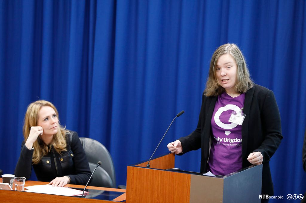 Marthe Øvrum fra Skeiv ungdom taler på et møte som barne- og likestillingsminister Linda Hofstad Helleland har invitert til. Ministeren sitter ved siden av Marthe og ser på henne. Dato for møtet var 31. januar 2018. Foto.