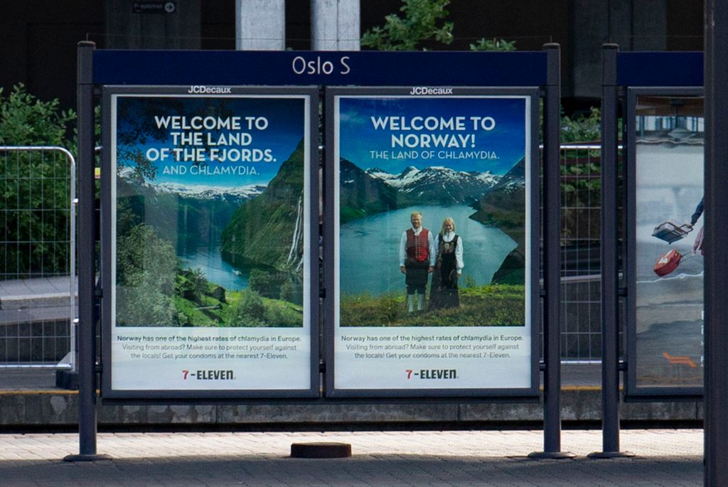 To reklameplakatar for 7-Eleven er sette opp på ei togplattform på Oslo S. Dei består av flotte bilete av fjord, fjell og eit blondt par i bunad og ein tekst som seier "Welcome to Norway – the land of Chlamydia". Foto.