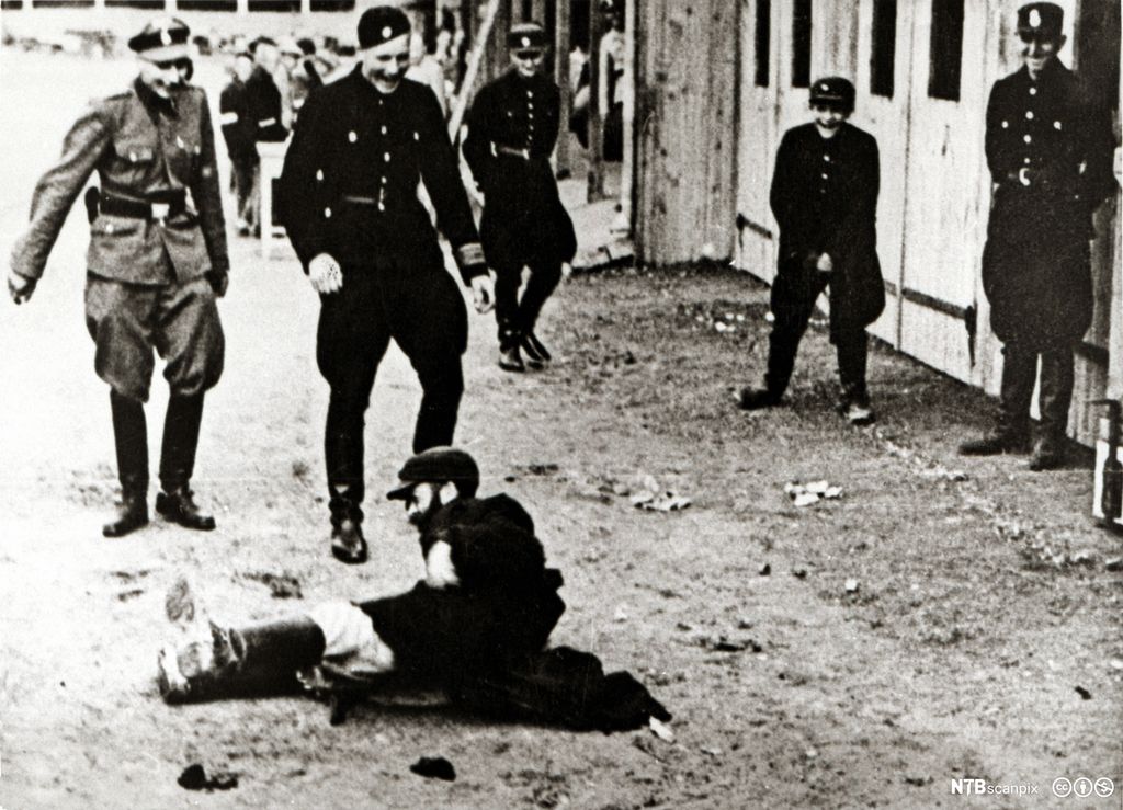 Vakter i uniform står rundt en mann som ligger på bakken. Vaktene ler. Foto.