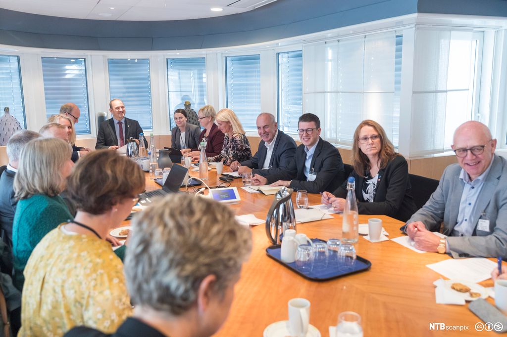 Vaksne i ulik alder sit rundt eit ovalt bord i eit stort og lyst møterom. Kanner, vasskaraflar, koppar og glas står midt på bordet. Foto.