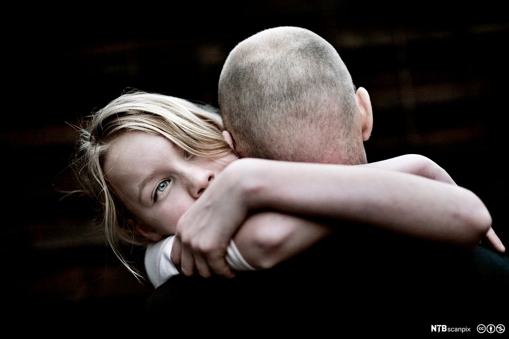 Far og datter holder rundt hverandre. Faren med snauklipt hode, og med ryggen til. Dattera med ansiktet mot betrakteren. Hun har lyst hår og blå øyne og ser alvorlig ut. Foto.
