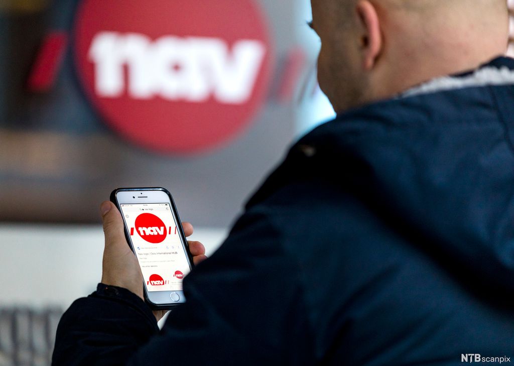 En mann holder en mobiltelefon med NAV-app utenfor et NAV-kontor. Foto.