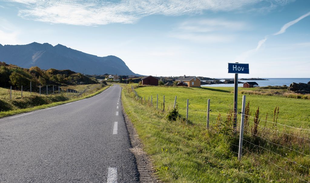 Smal asfaltvei og skilt med stedsnavnet Hov. Høye fjell og hav i horisonten, jorder på hver side av veien. Sol og blå himmel. Foto.