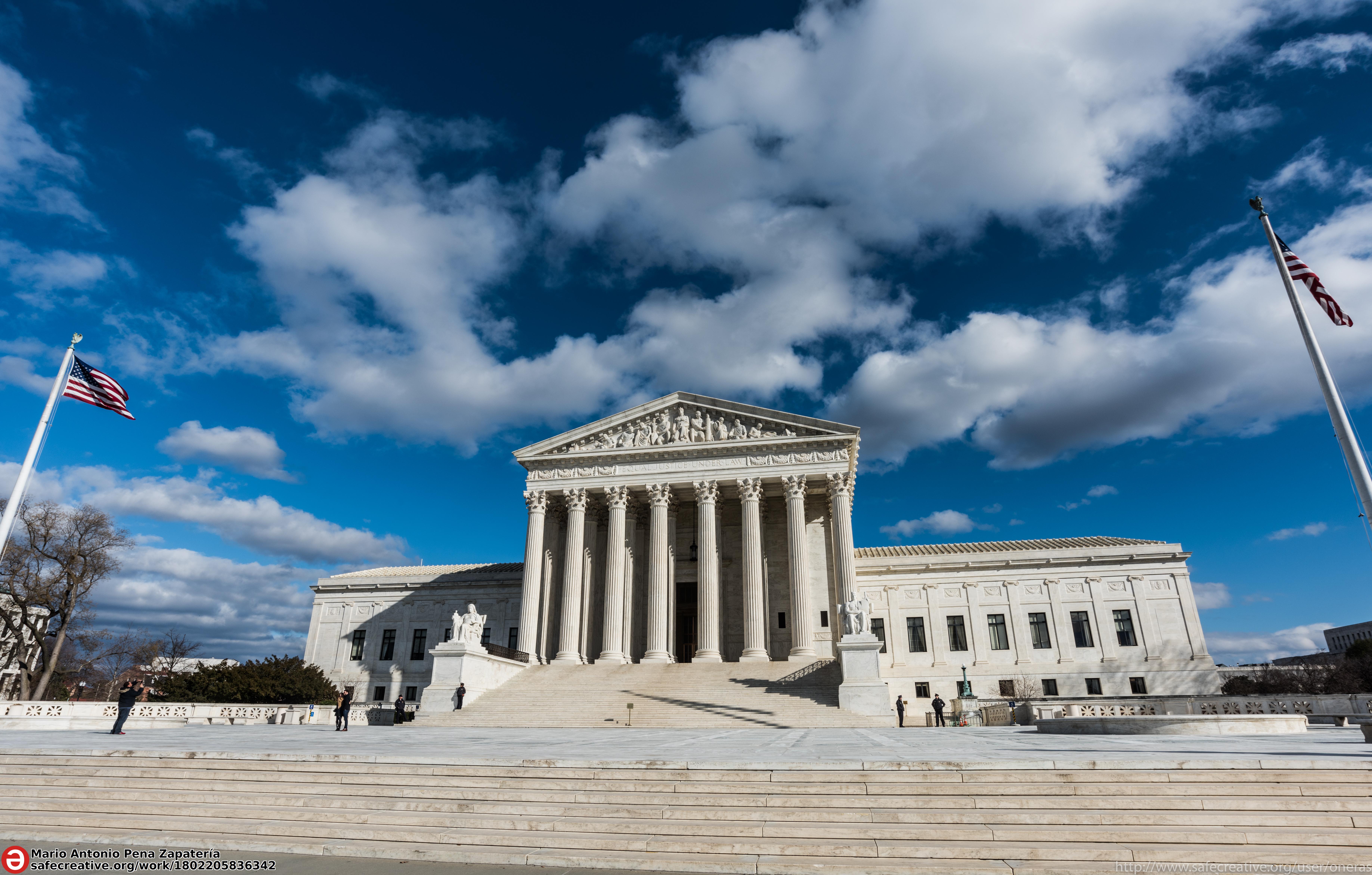 Photo: We see a large white building with white steps leading up to a structure that looks like a temple from ancient Greece, with columns and a pediment decorated by a frieze. Behind this structure, we see that the rest the building has more of a square shape, with many windows in rows.  