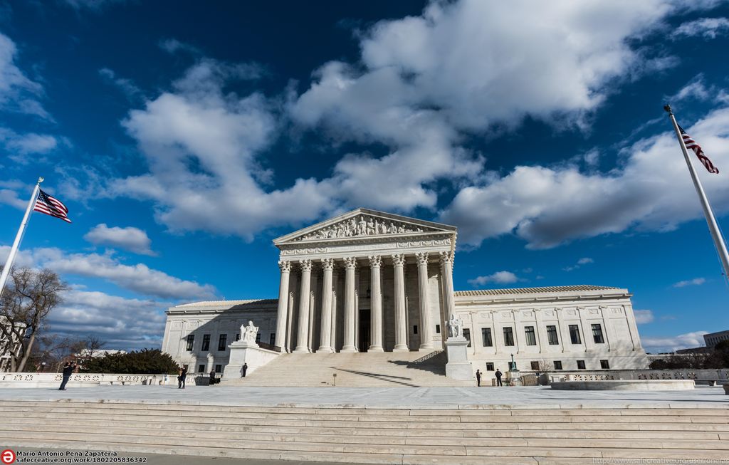 A large white building with white steps leading up to a structure that looks like a temple from ancient Greece, with columns and a pediment decorated by a frieze. Behind this structure, the rest of the building has more of a square shape, with many windows in rows. Photo.