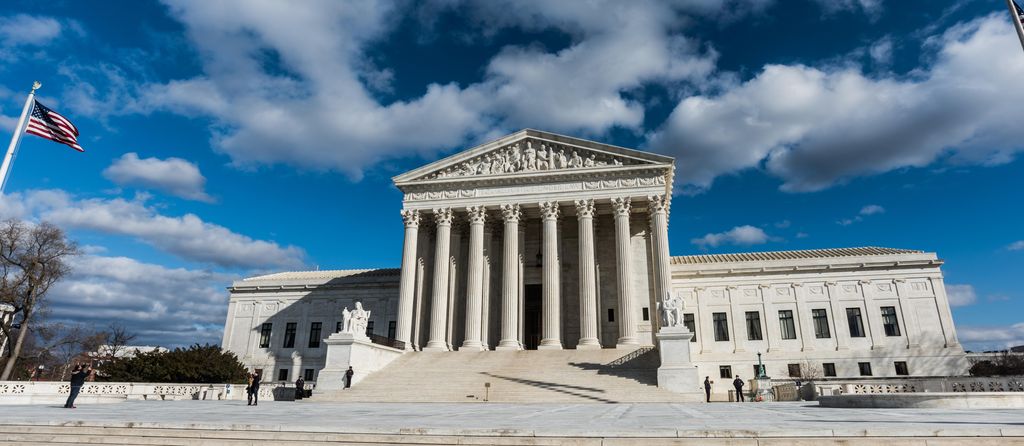 Photo: We see a large white building with white steps leading up to a structure that looks like a temple from ancient Greece, with columns and a pediment decorated by a frieze. Behind this structure, we see that the rest the building has more of a square shape, with many windows in rows.