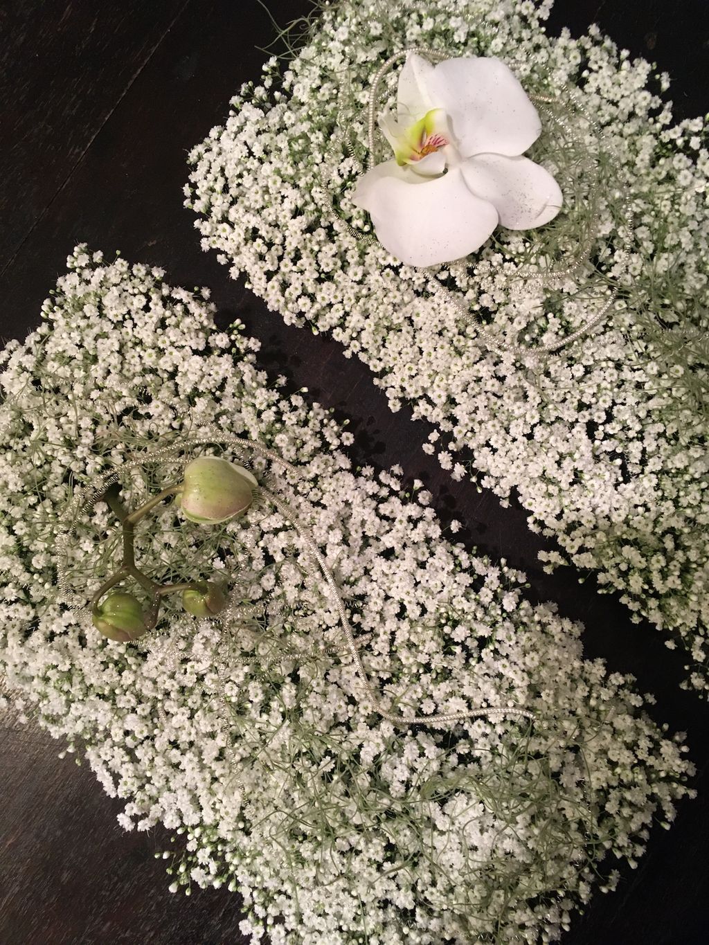 To skråstilte, stor flater fylt med veldig små, hvite blomster. Oppå den ene flaten ligger det en hvit orkidé, og oppå den andre flaten ligger det noen orkidé-blomsterknopper. Bakgrunnen er mørk. Foto.