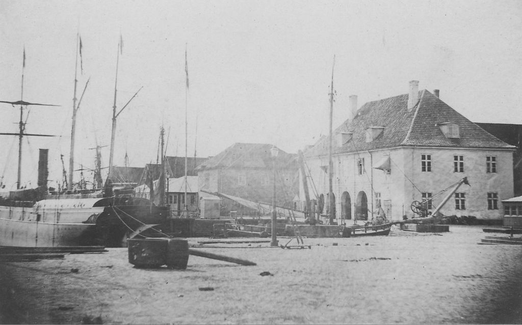 Tollboden i Christiania med flere bygg langs bryggen og båter til kai. Foto i svart-hvitt.