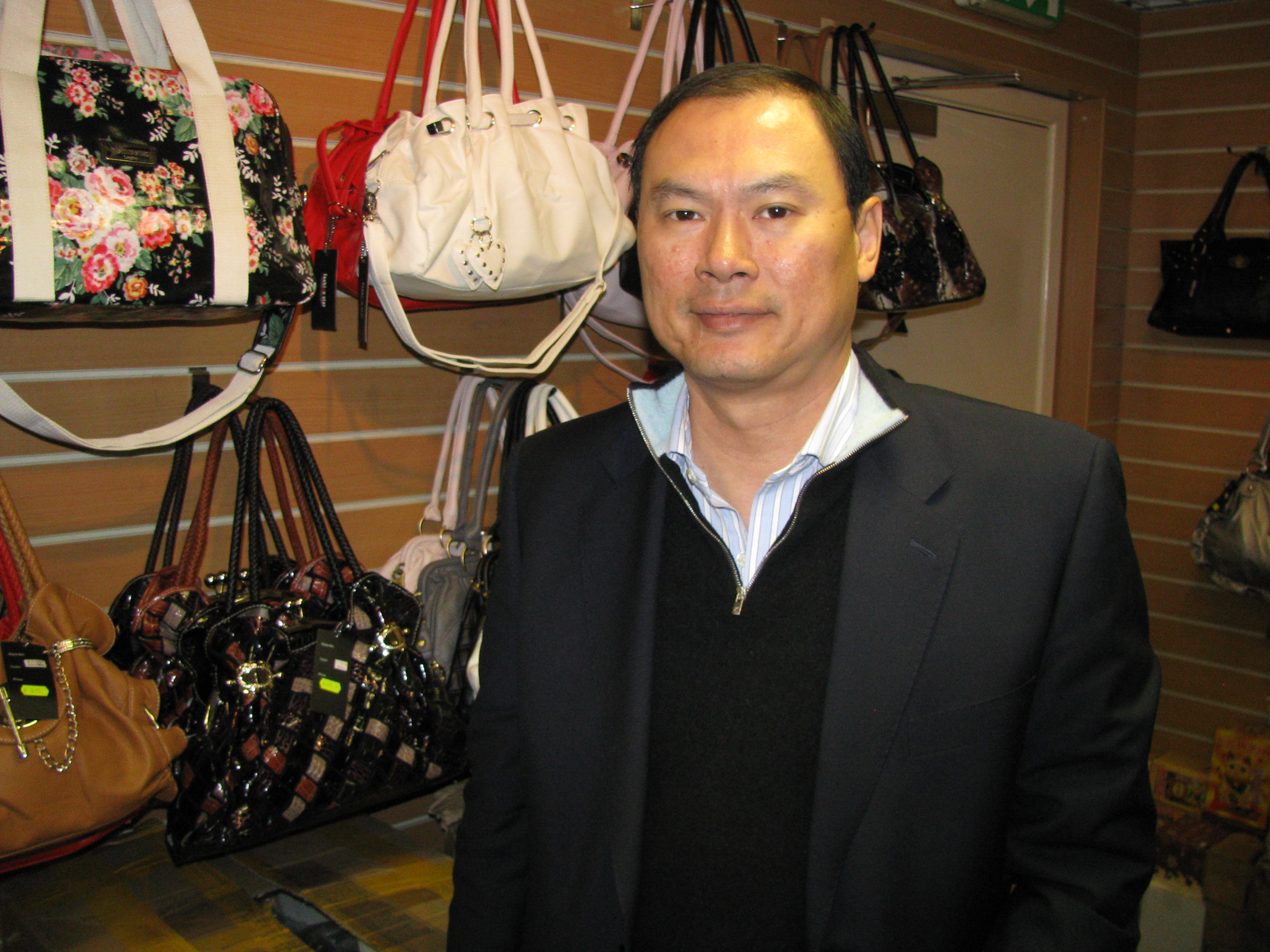 Adult Asian man who is wearing a suit jacket, sweater, and open shirt. He is looking at the camera. He appears to be in a shop that sells handbags. Photo.