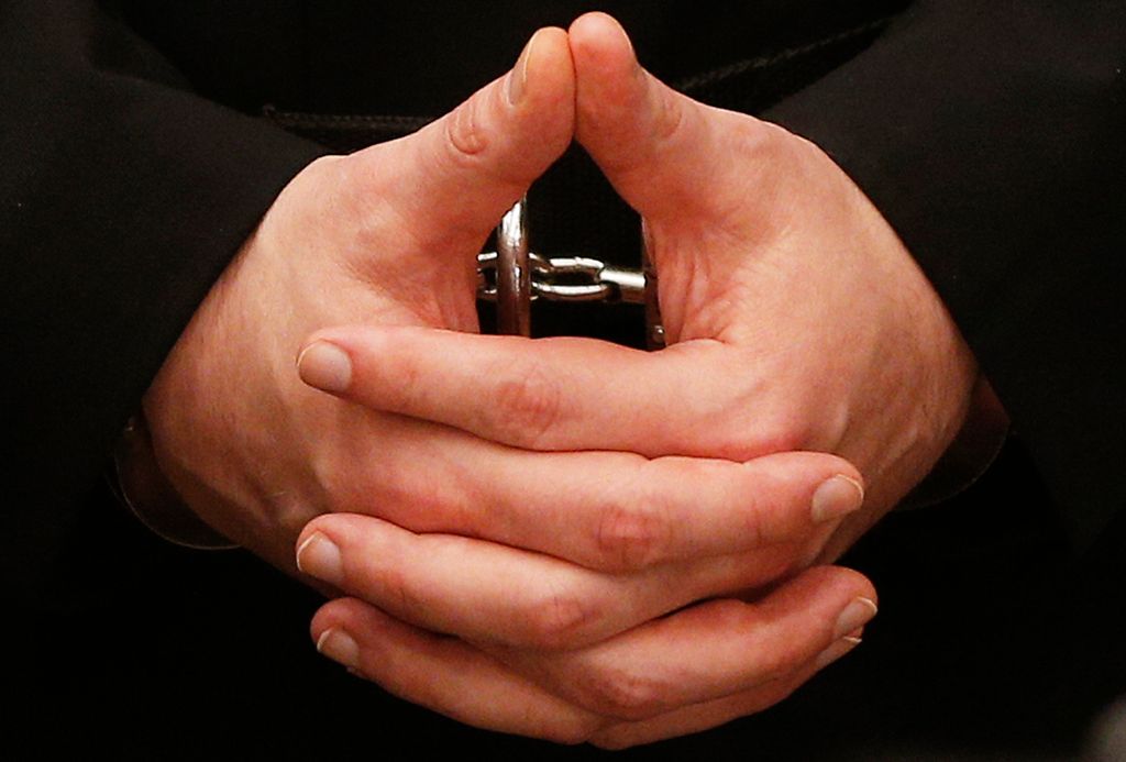 Nærbilete av falda mannehender i handjern. Fingrane er korte og kraftige, neglene er kortklipte. Foto.