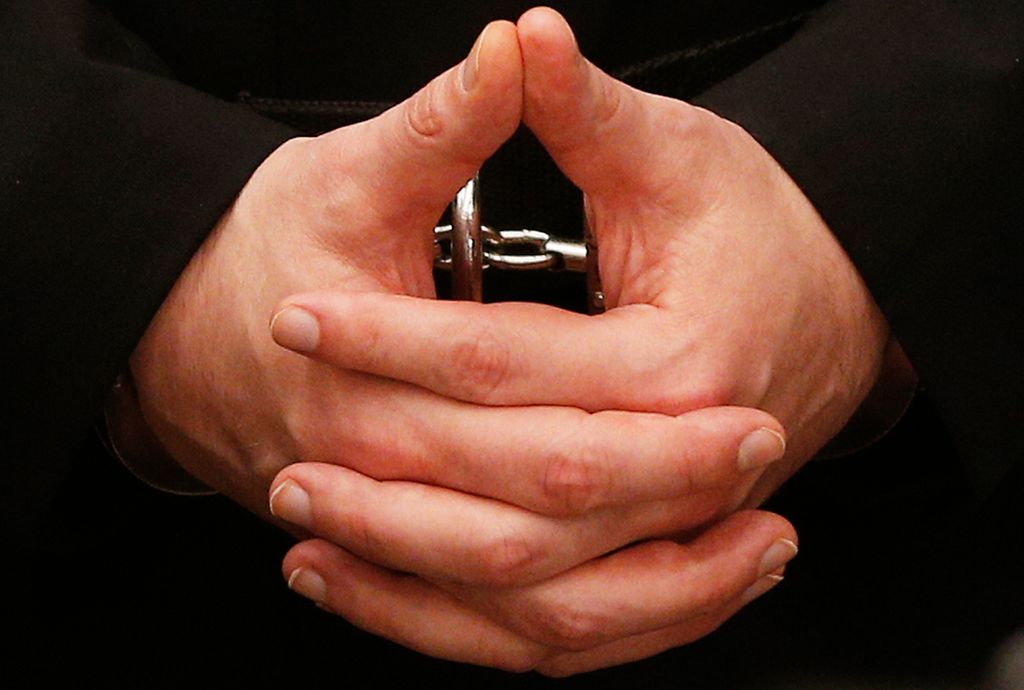 Nærbilde av foldede mannehender i håndjern. Fingrene er korte og kraftige, neglene er kortklipte. Foto.