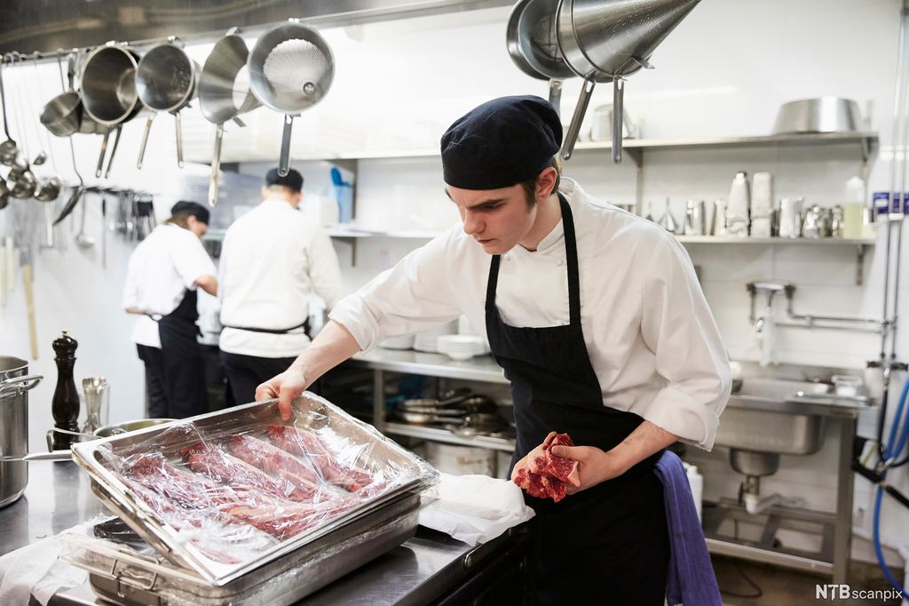 En kokk på et kjøkken undersøker kjøttstykker i en bakke. Foto.