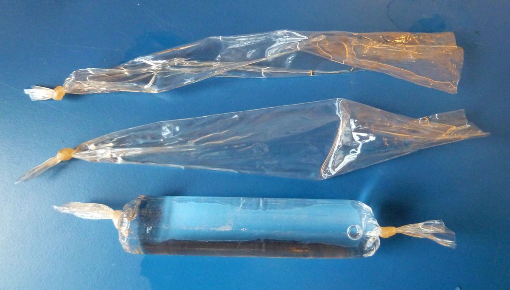 Tre avklipte bitar av ein tynn plastslange ligg på eit bord. To av dei er tomme og knytte i den eine enden, mens den tredje er knytt i begge endar og fylt med ei gjennomsiktig væske slik at han liknar ei pølse. Bilete.