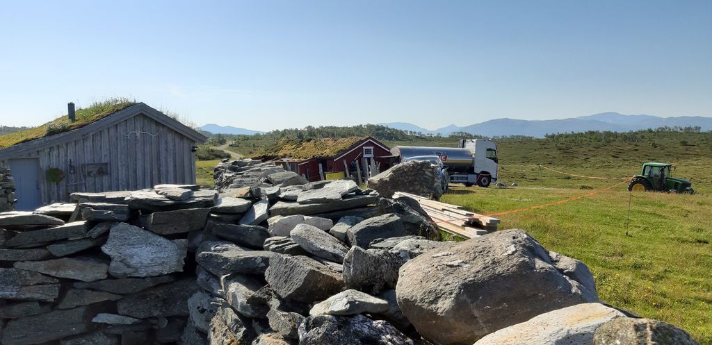 Et seterlandskap med en steinmur i forgrunnen. I bakgrunnen er det to bygninger, og det står en tankbil med TINE-logo ved den ene. Det står også en traktor på graset til høyre lenger bak, og det er fjell i horisonten. Foto.