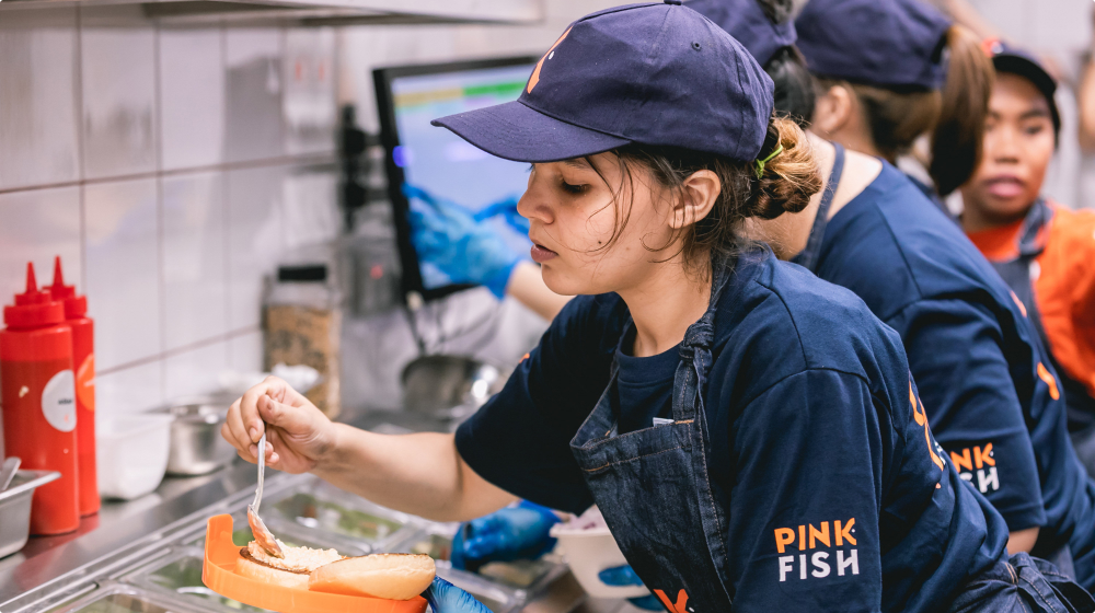 En ansatt i mørkeblå t-skjorte, kaps og dongery-forkle ordner med en lakseburger på et storkjøkken. Foto.