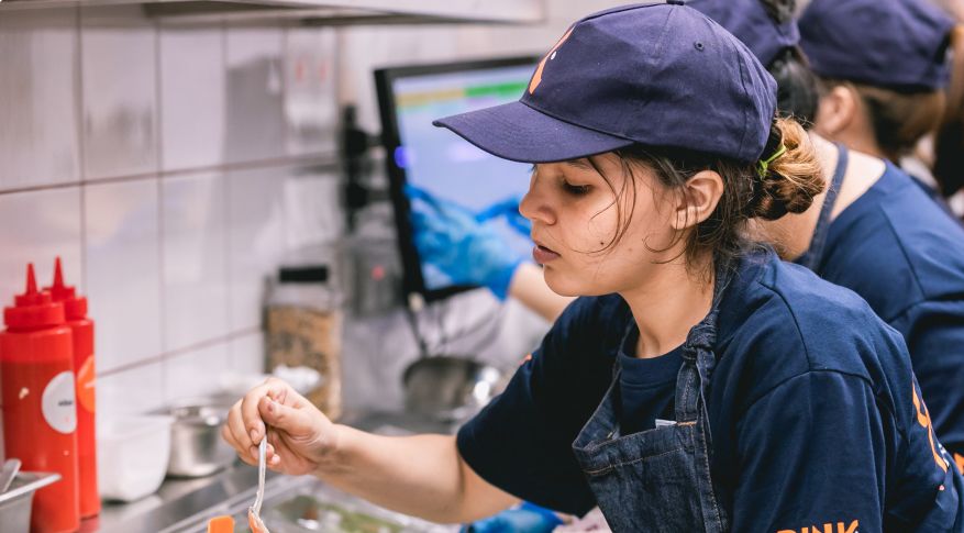 En ansatt i mørkeblå t-skjorte, kaps og dongery-forkle ordner med en lakseburger på et storkjøkken. Foto.
