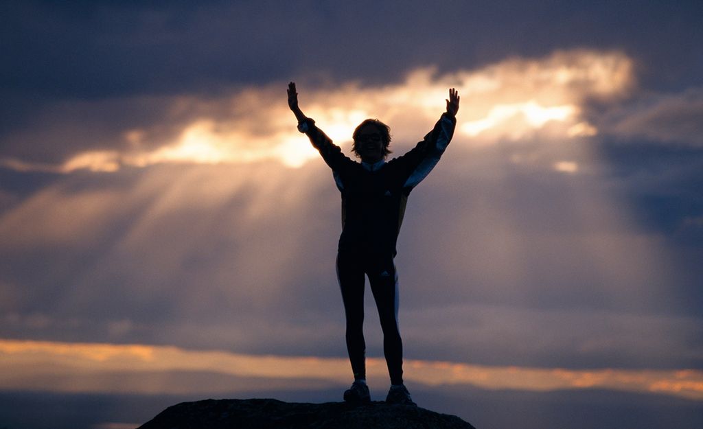 En person strekker armene opp mot en himmel der sola bryter gjennom skyene. Foto.