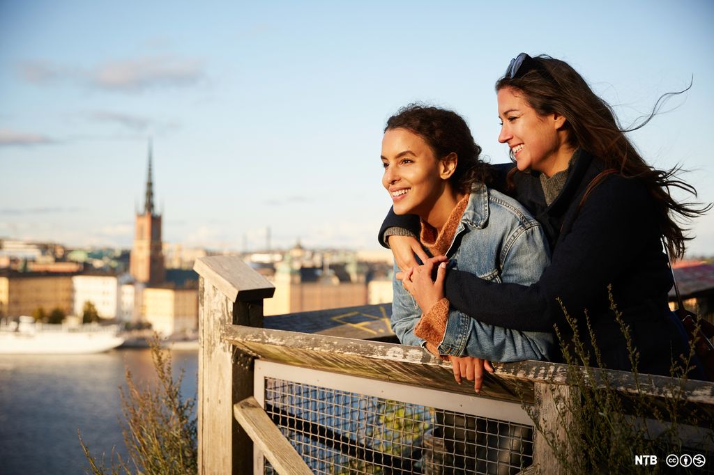 To kvinner står og holder rundt hverandre, mens de ser ut over en by. Foto.