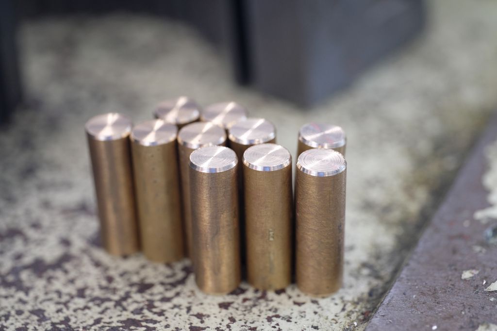 Ti dreide brikker i kobberfarga metall. Brikkene har fas. Foto.