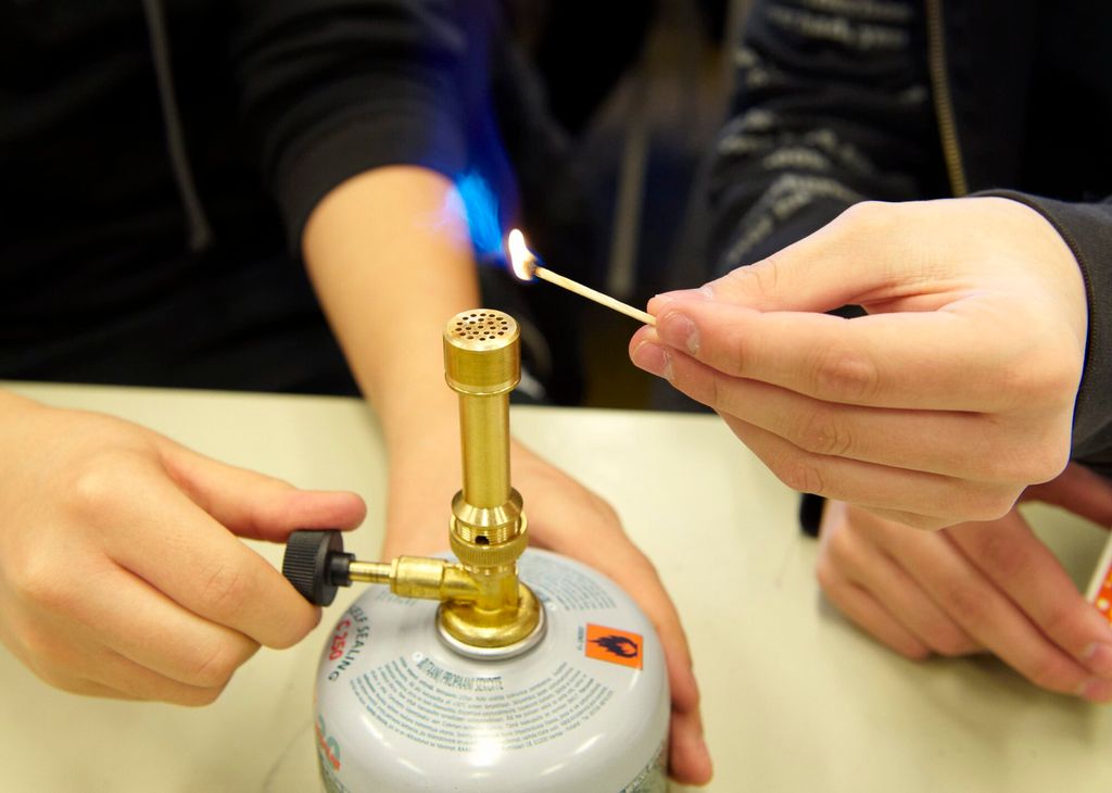 En person skrur på gassventilen, og en annen holder en tent fyrstikk mot brennerhodet på en gassbrenner. Foto.