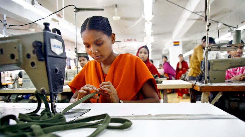 Ungjente som sitter ved en symaskin på en klesfabrikk i Bangladesh. I bakgrunnen er flere tekstilarbeidere som sitter ved symaskinene, alle er unge og har fargerike klær. Foto.