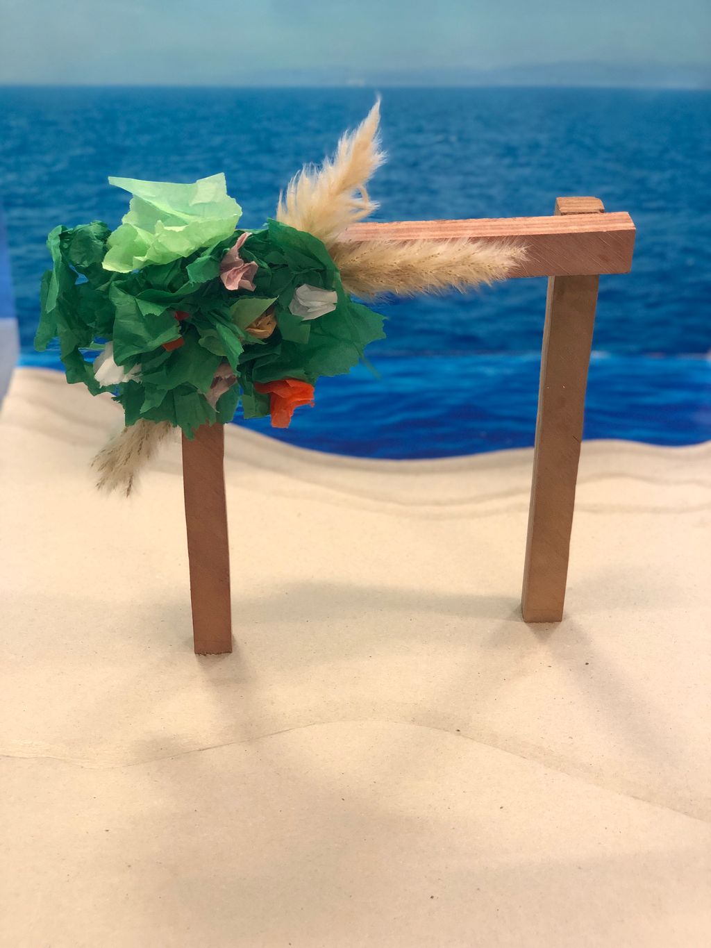 Prototype av bryllupsportal i tre med pampas, grønt og blomster plassert i en modell av strand. Foto.