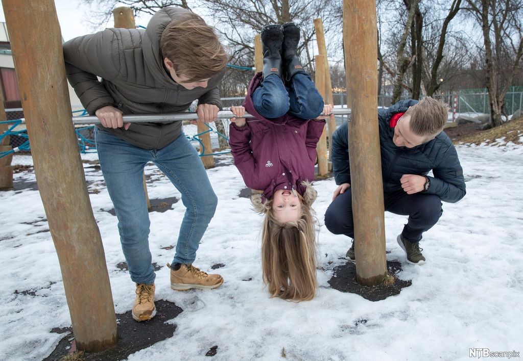 Ei jente heng opp ned i eit klatrestativ, håret hennar heng ned i bakken. To unge menn står ved sida av ho og ser på i leiken, den eine sit på huk og den andre bøyer seg over stonga ho snurrar rundt. Det er snø på bakken. Foto.