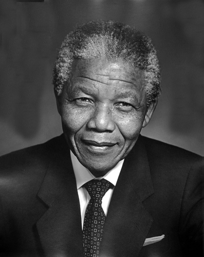 A black and white photo of Nelson Mandela. He's dressed in a suit and tie. He looks directly into the camera, smiling.  