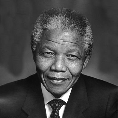 A black and white photo of Nelson Mandela. He's dressed in a suit and tie. He looks directly into the camera, smiling.