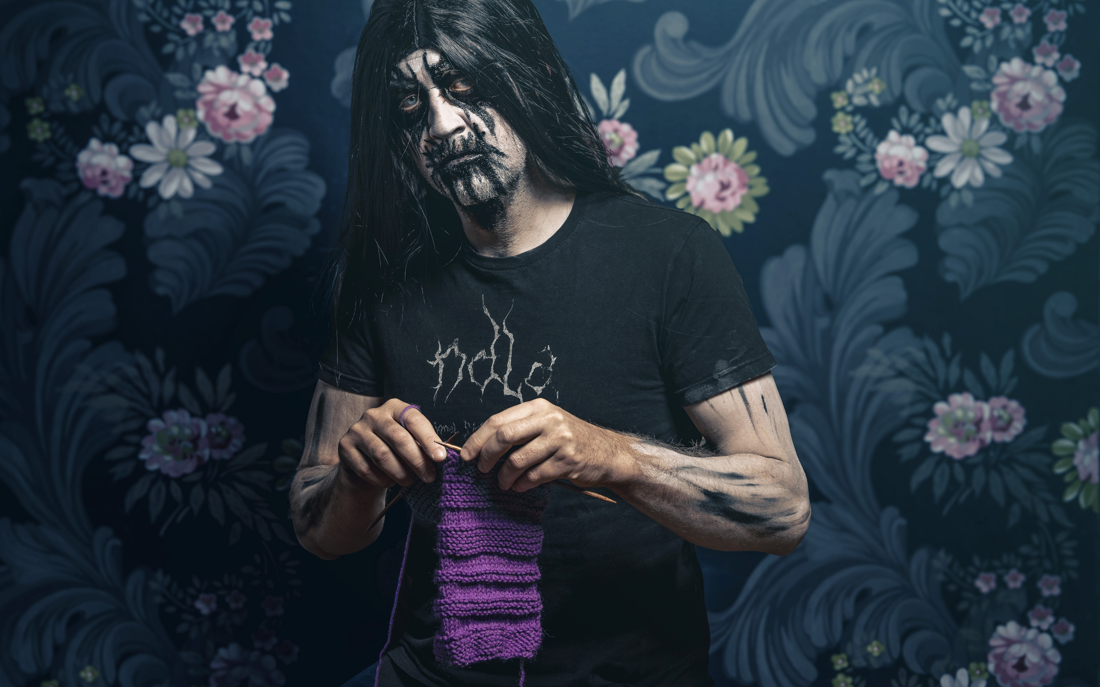 Mann med blackmetal-liksminke strikker et skjerf i fargen lilla. Han står i et rom med blomstret tapet. Foto.