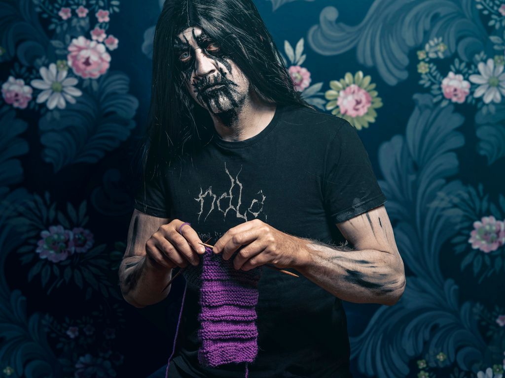 Mann med blackmetal-liksminke strikker et skjerf i fargen lilla. Han står i et rom med blomstret tapet. Foto.