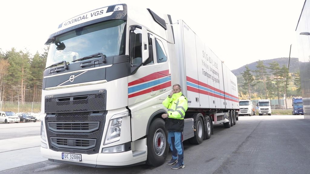 En yrkessjåfør i refleksjakke står ved siden av et hvitt vogntog og gjør tommel-opp-tegn. Foto.