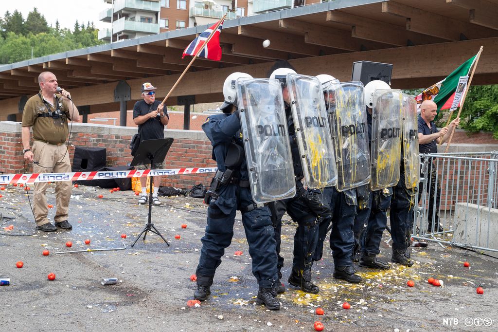 En middelaldrende mann med mikrofon og en eldre mann med norsk flagg står bak en politisperring. Foran står det flere politifolk med hjelmer og skjold. Foto.