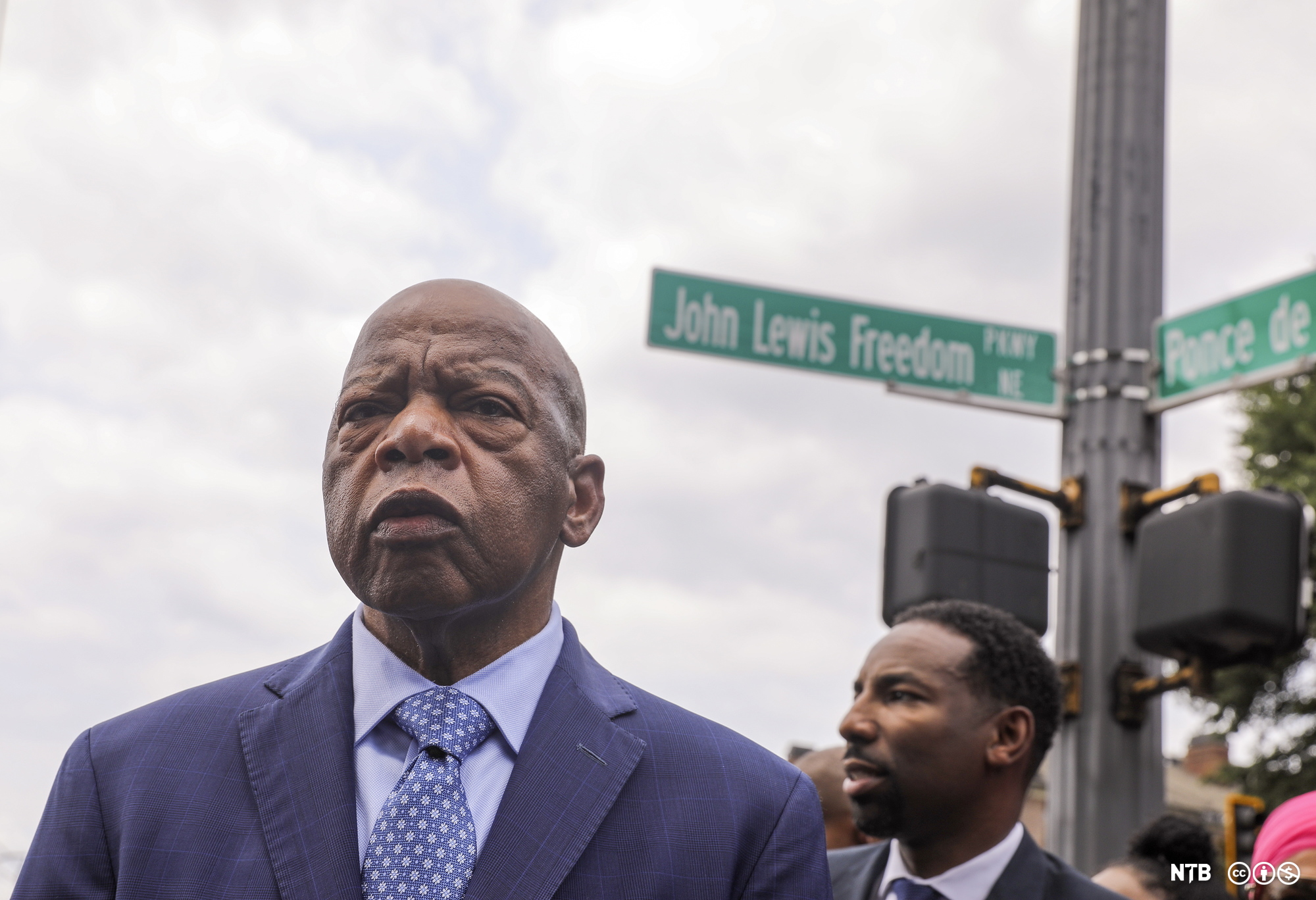 Photo: We see John Lewis, he is an older Black man. He is bald. He is wearing a blue suit with a tie. Behind him we see a younger man. There is a street sign behind them. The sign says John Lewis Freedom. 