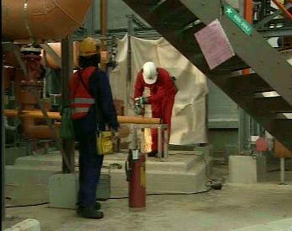 En arbeider med hjelm og rød refleksvest ser på at en annen arbeider kutter rør med håndverktøy så gnistene spruter. Foto.