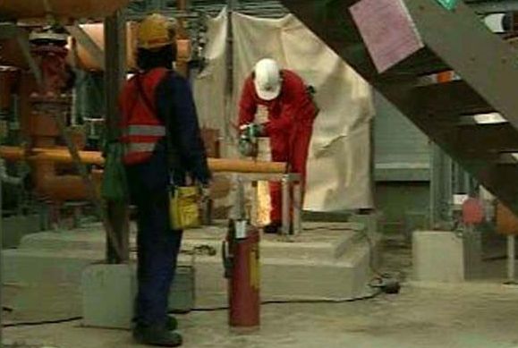 En arbeider med hjelm og rød refleksvest ser på at en annen arbeider kutter rør med håndverktøy så gnistene spruter. Foto.