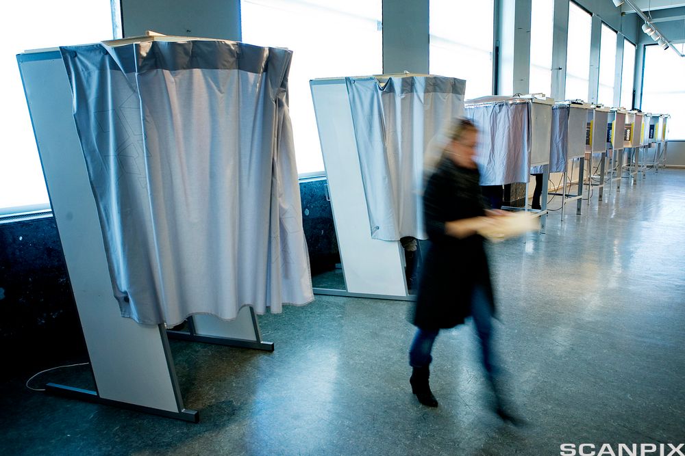 Flere stemmeavlukker står på rekke i et stemmelokale. En kvinne er på vei bort fra et stemmeavlukke med en stemmeseddel i handa. Foto.