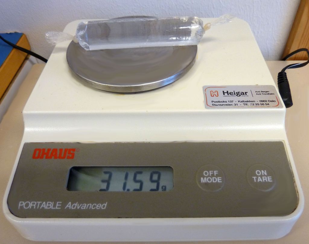 Ein gjennomsiktig bit av ein plastslange er knytt som ei pølse og fylt med vatn og lagt på ei lita vekt. Vekta viser 31,59 gram. Bilete.