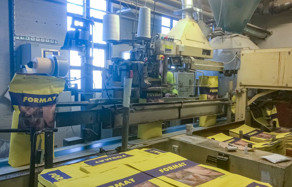 Produksjonslinje i en kraftfôrfabrikk. Det ligger mange papirsekker klare til å fylles med kraftfôr foran en maskin som plukker opp sekkene, fyller dem med kraftfôr og lukker dem ved å sy dem sammen i toppen. Foto.