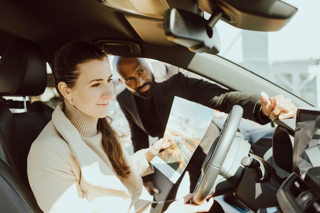En kvinne sitter i førersetet på en parkert bil med en brosjyre i hånda. En mannlig selger veileder og forklarer om bilen. Foto.